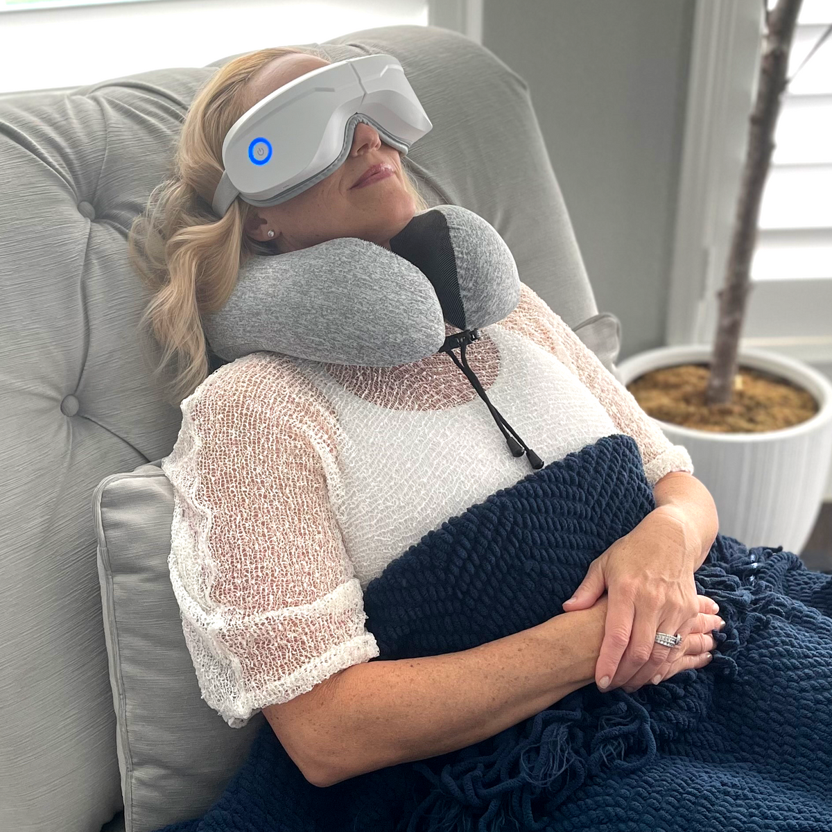 Woman resting in a chair wearing the Relaxable eye massager and a gray travel neck pillow
