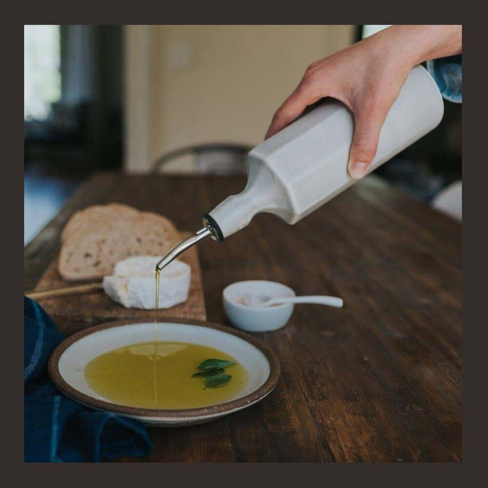 Handcrafted porcelain olive oil dispenser pouring olive oil onto a plate with bread and cheese on a wooden table.