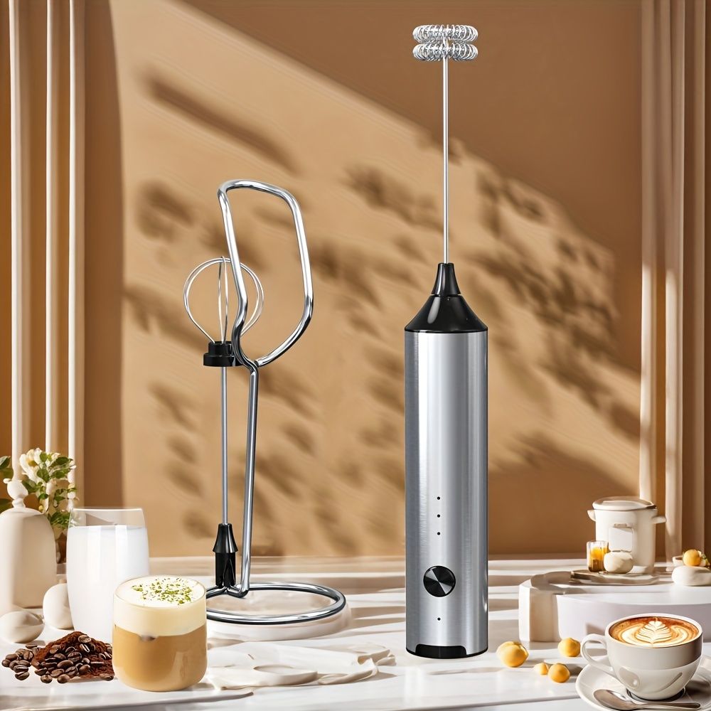 Rechargeable handheld milk frother in silver with dual whisk heads and two frothing attachments displayed with coffee drinks